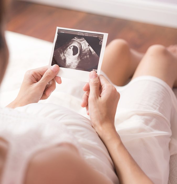 Pregnant asian woman looking at ultrasound scan of baby and touching her belly, close up of scan